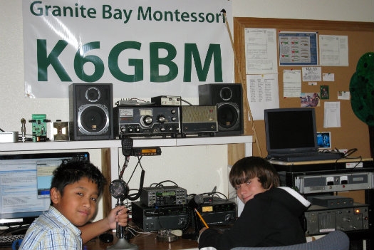 Two students looking at the camera at their radio setup with a green K6GBM Granite Bay Montessori school banner behind them on the wall.